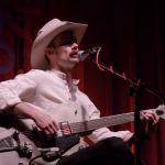 A man in a western hat sits playing a white electric guitar and sings.