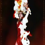 A woman wearing a red dress, with a white guitar beside her, is seen on fire, raising an arm to the sky