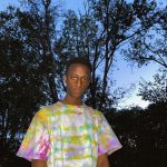 Ultravioletenvy, wearing a tie-dyed T-shirt, stands in the woods facing the camera