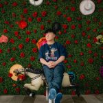 Andy Yu, wearing jeans and a T-shirt, sits in front of a green wall dotted with flowers